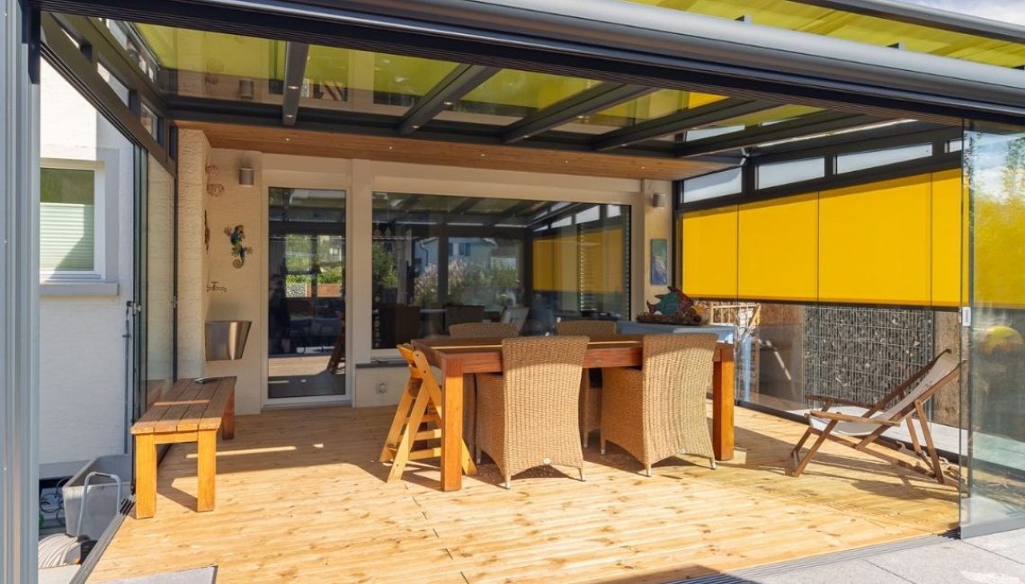 Wintergarten mit Glasdach und gelben Sonnenschutz-Elementen, ausgestattet mit einem Esstisch und Rattan-Stühlen auf Holzboden.