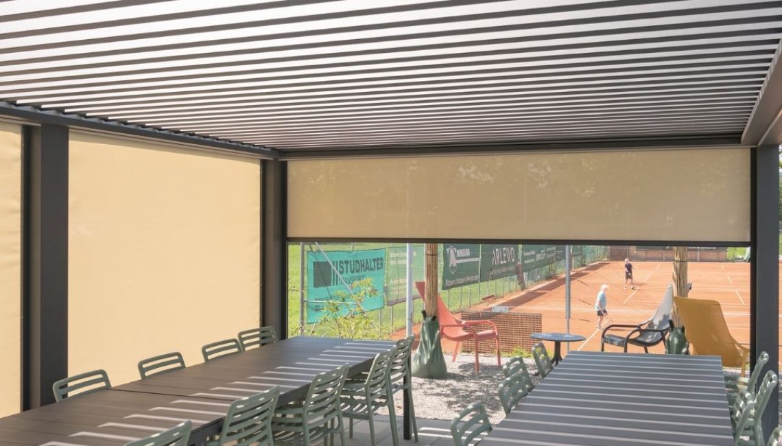 Innenansicht des Lamellendachs beim Tennisclub Horw in Luzern mit geschlossenen Seitenmarkisen, langen Tischen und Blick auf die Tennisplätze.