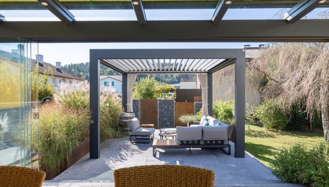 Blick aus dem Wintergarten mit Glasdach auf ein modernes Lamellendach mit Lounge im Garten.