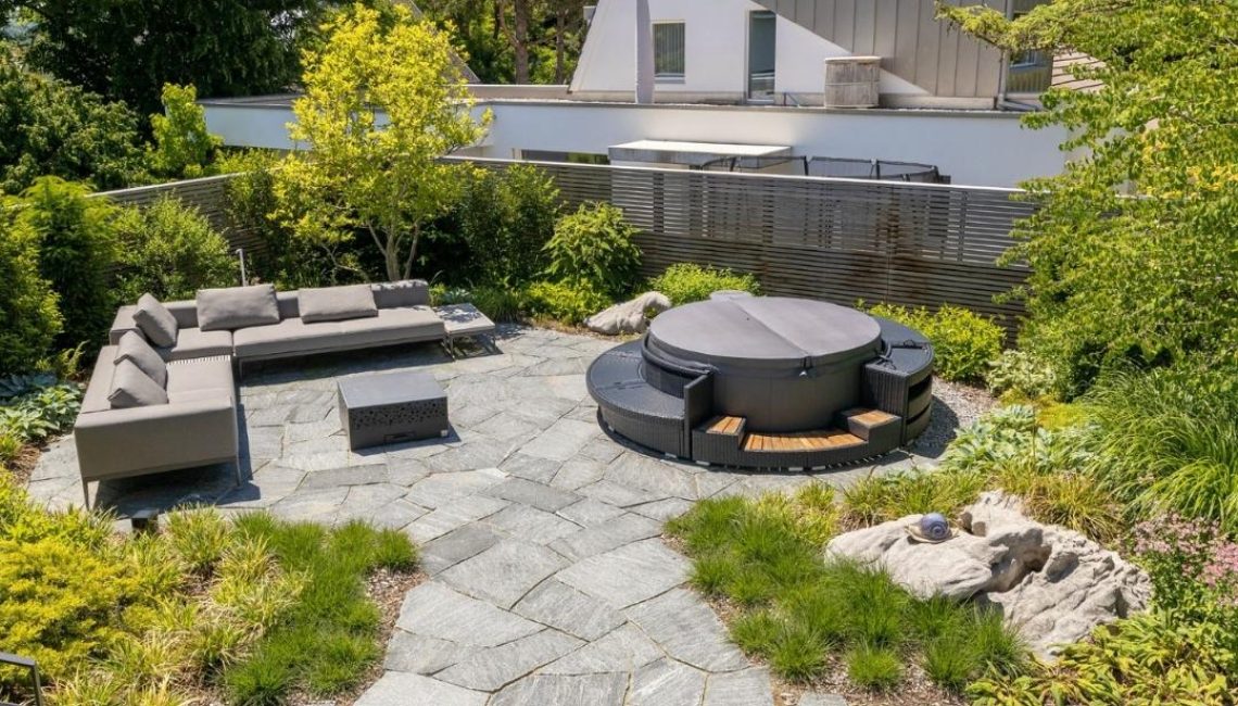Moderner Gartenbereich mit Natursteinboden, gemütlicher Lounge, Whirlpool und dichter Bepflanzung für ein harmonisches Ambiente.