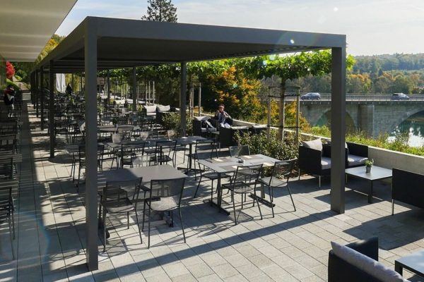 SKL 100 Lamellendach auf einer grosszügigen Restaurantterrasse mit Blick auf die Brücke und den Fluss.