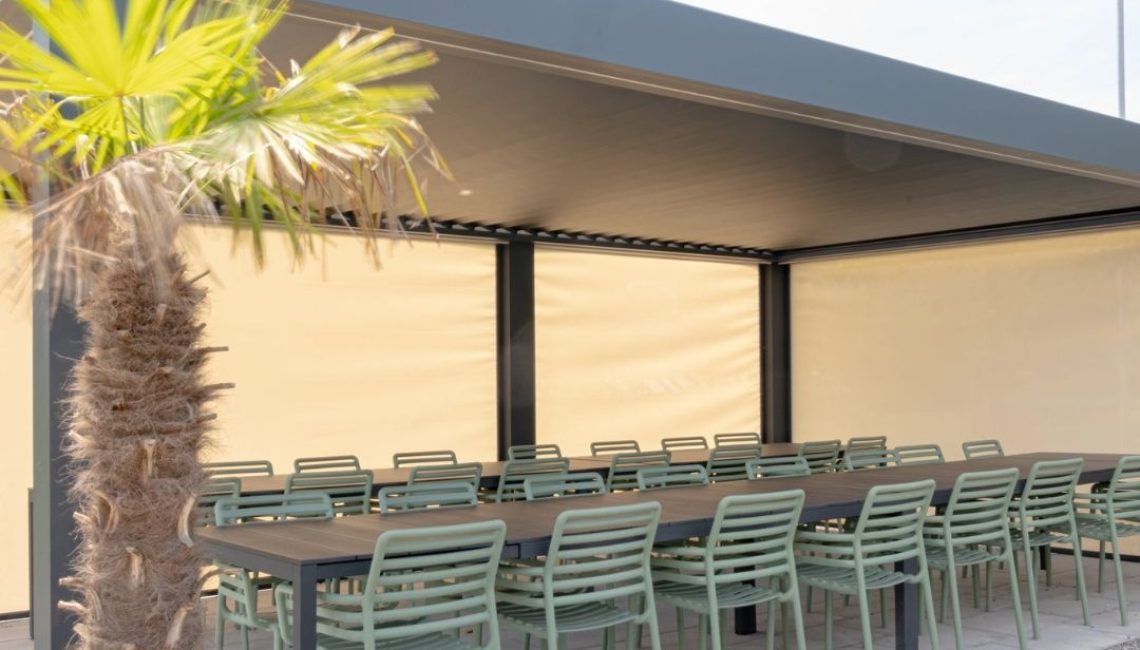 Lamellendach beim Tennisclub Horw in Luzern – überdachter Sitzbereich mit langen Tischen, grünen Stühlen und geschlossenen Senkrechtmarkisen, Palmenpflanze im Vordergrund.