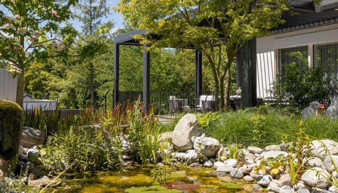 Moderner Garten mit Naturteich, Seerosen und grossen Steinen, im Hintergrund ein anthrazitfarbenes Lamellendach mit gemütlichem Sitzbereich.