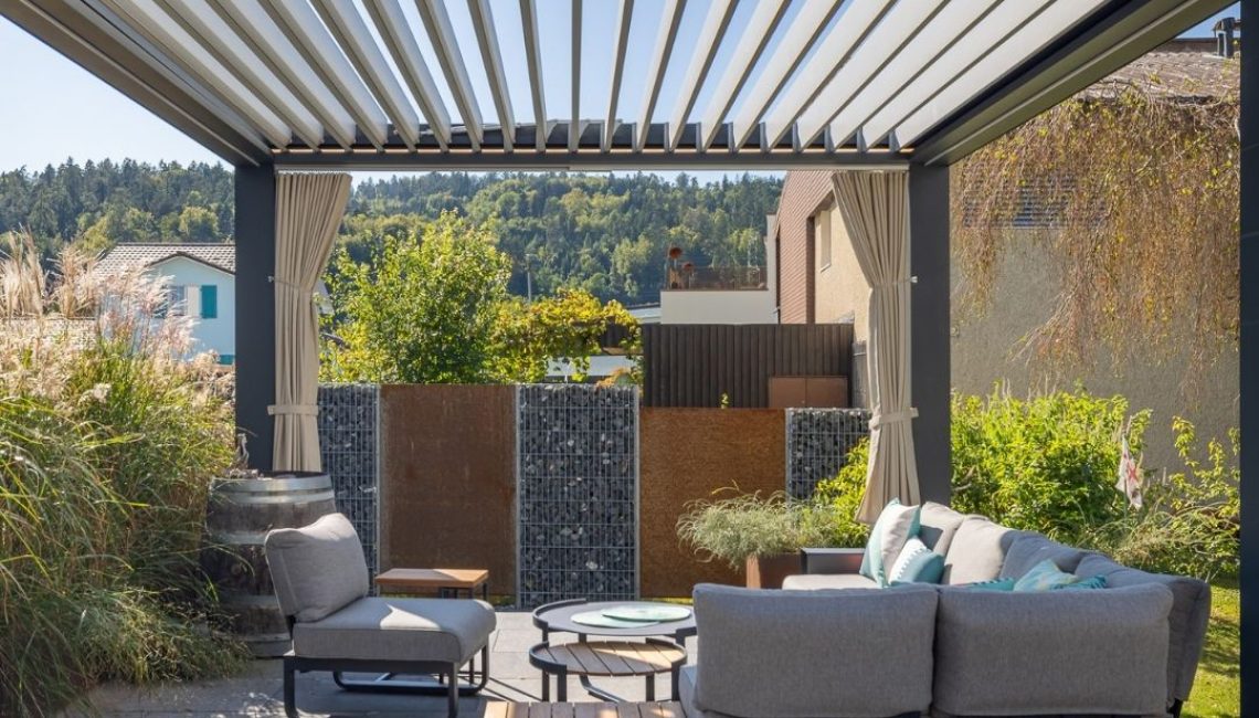 Offenes Lamellendach mit moderner Outdoor-Lounge darunter, umgeben von Grünpflanzen und Sichtschutzwänden.