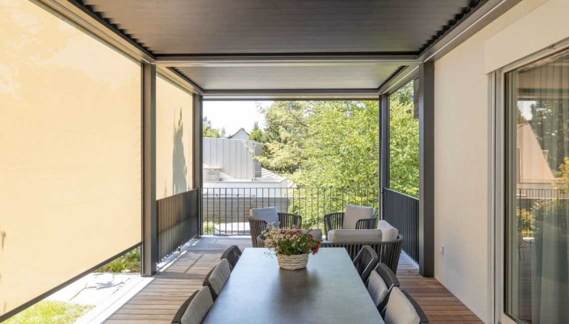 Überdachter Essbereich mit grossem Tisch und Stühlen unter einem anthrazitfarbenen Lamellendach, seitliche Screens sorgen für Sicht- und Sonnenschutz, Blick in den Garten.