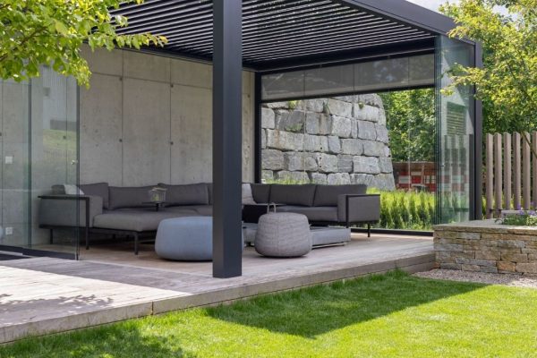 SKL 100 Lamellendach mit Glaswänden und moderner Lounge-Einrichtung in einem Garten.