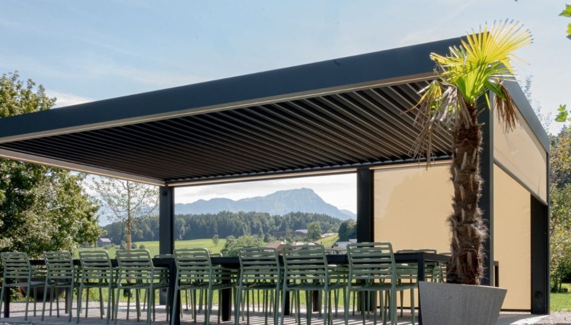 Lamellendach beim Tennisclub Horw in Luzern – überdachter Sitzbereich mit langen Tischen, Palmenpflanze im Vordergrund und Bergpanorama im Hintergrund.