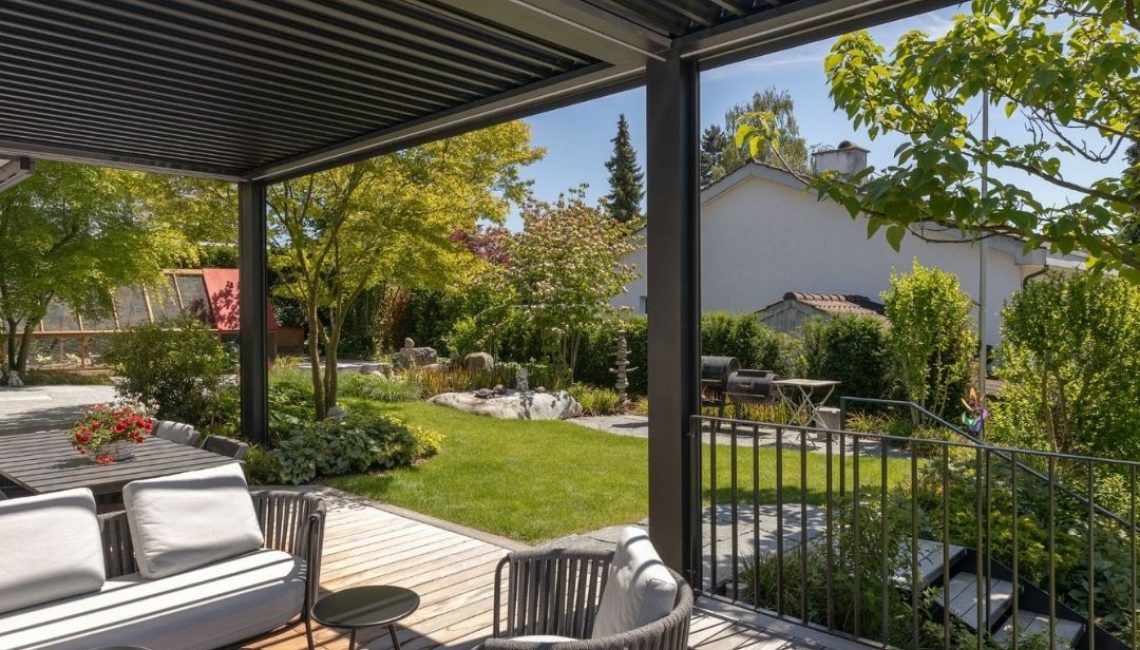 Gemütlicher Sitzplatz mit Lounge-Möbeln unter einem anthrazitfarbenen Lamellendach, Blick in den gepflegten Garten mit Rasen, Bäumen und Naturstein-Elementen, Metallgeländer und Holzdeck im Vordergrund.