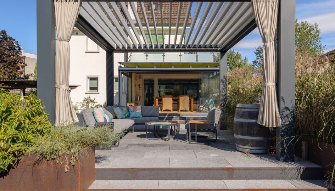 Frontansicht eines modernen Lamellendachs mit offener Lamellenstellung, darunter eine gemütliche Lounge mit Sofas und Couchtischen, im Hintergrund ein verglaster Wintergarten.