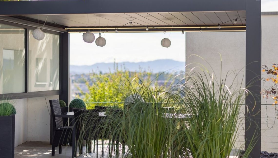 SKL 100 Lamellendach mit seitlicher Verglasung – moderner Essplatz im Freien, dekoriert mit Pflanzen und Laternen, im Hintergrund Bergpanorama.