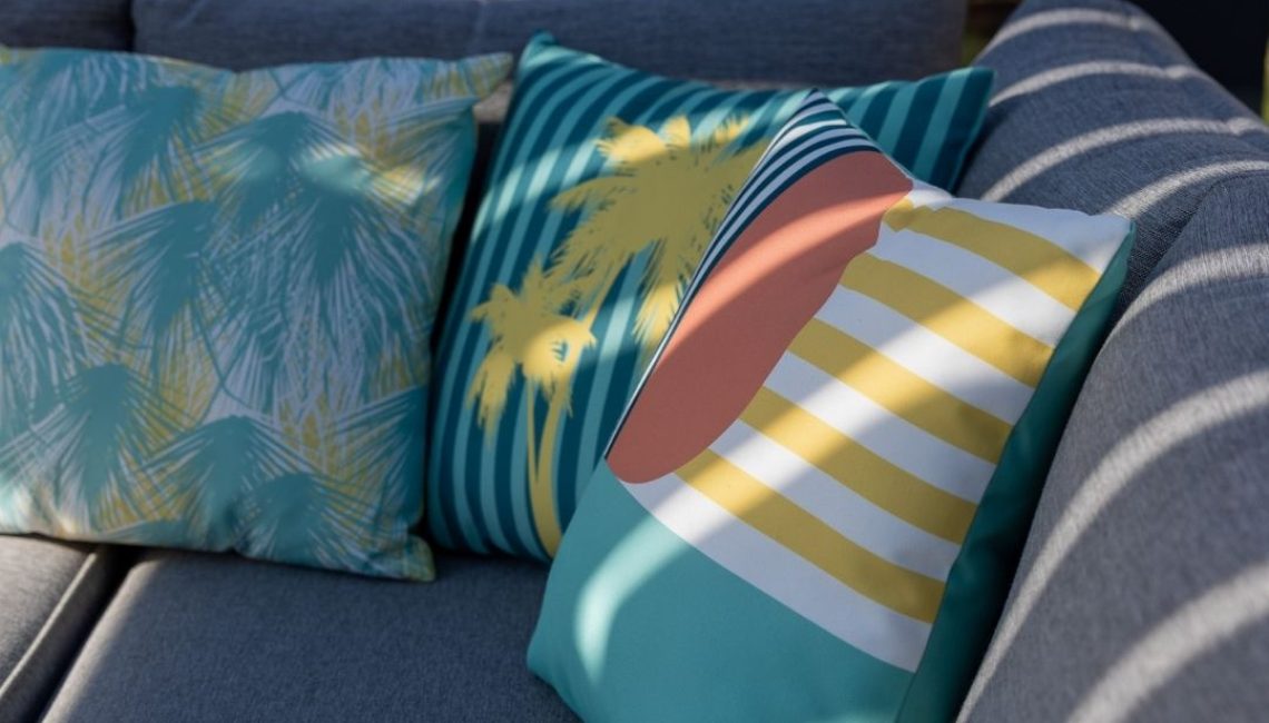 Nahaufnahme bunter Dekokissen auf einem grauen Outdoor-Sofa, mit Schattenwurf der Lamellen eines Lamellendachs.