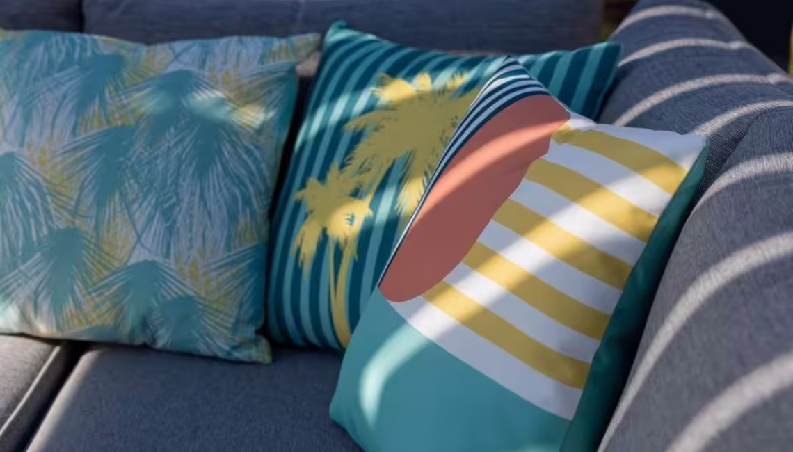 Nahaufnahme bunter Dekokissen auf einem grauen Outdoor-Sofa, mit Schattenwurf der Lamellen eines Lamellendachs.