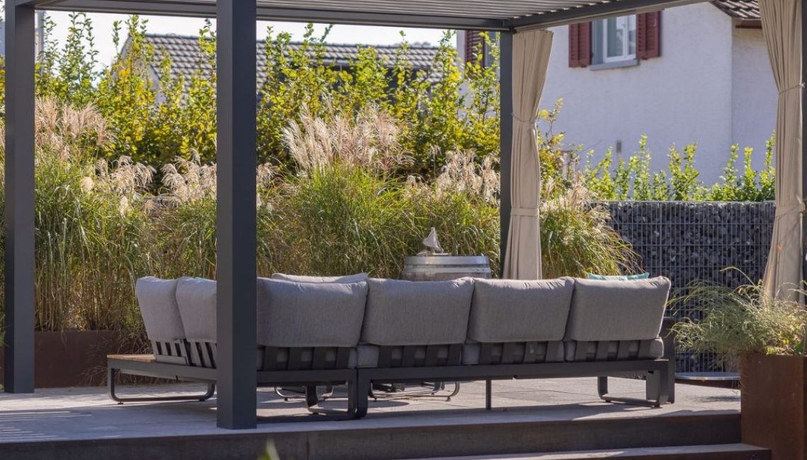 Seitliche Ansicht eines modernen Lamellendachs mit grauem Outdoor-Sofa, eingerahmt von hohen Gräsern und einer Natursteinwand im Hintergrund.