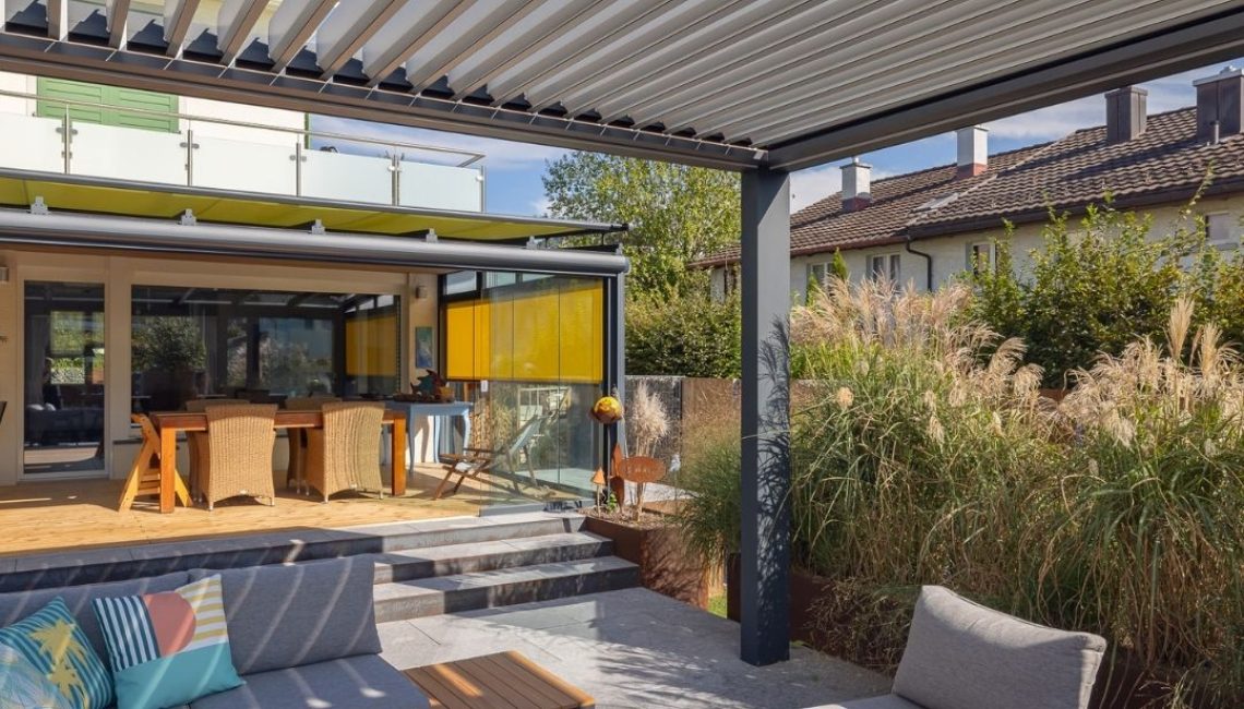Blick unter ein modernes Lamellendach mit gemütlicher Lounge und angrenzendem Wintergarten mit gelben Sonnenschutz-Elementen.