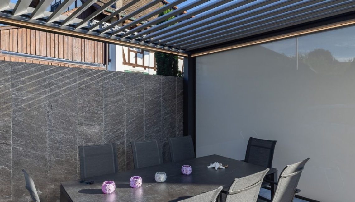 Essbereich unter einem Lamellendach mit seitlichen Screens und Sichtschutzwand aus Natursteinoptik.