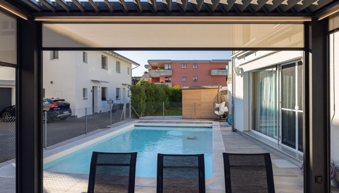 Blick vom gedeckten Essbereich unter einem Lamellendach auf den Pool im Garten.