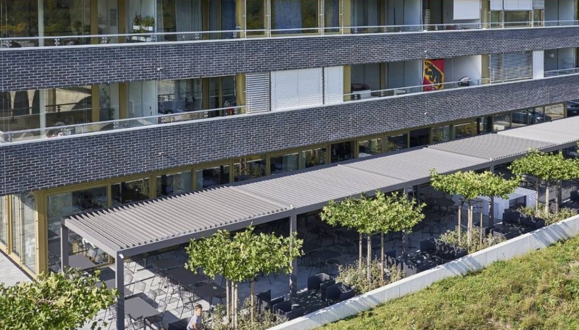 Grossflächiges SKL 100 Lamellendach in Hinterkappelen Bern, integriert in die Architektur eines modernen Wohn- und Gastronomiegebäudes mit Terrasse und Sitzbereichen.