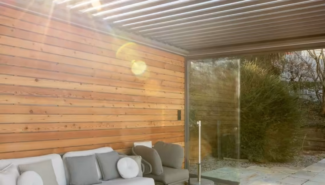 SKL 100 Lamellendach in Horgen, Zürich mit 300 kg/m² Schneelast – Sonnenstrahlen durch verstellbare Lamellen über einer gemütlichen Lounge mit Holzwand.
