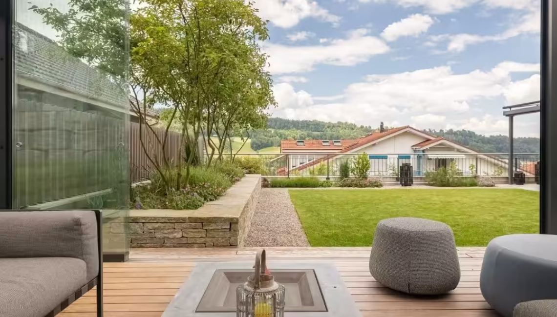 Blick aus einem SKL 100 Lamellendach mit Glaswänden in Malters, Luzern – moderner Outdoor-Bereich mit Lounge-Möbeln, Holzdeck und Blick ins Grüne.