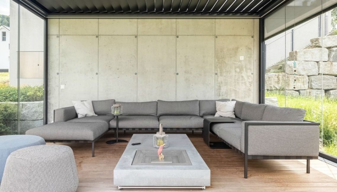 Innenansicht eines SKL 100 Lamellendachs in Malters, Luzern – geschlossene Lamellen, Glaswände und gemütliche Lounge-Einrichtung auf Holzdeck.