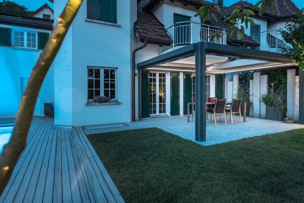 SKL 150 Panorama Plus Lamellendach bei Abendstimmung mit LED-Beleuchtung, vollständig zurückgefahrenen Lamellen und gedecktem Essbereich im Garten.