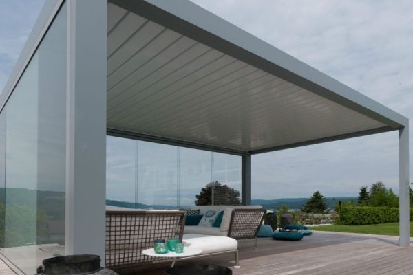 SKL 100 Lamellendach mit Glaswänden auf einer Holzterrasse mit Aussicht ins Grüne.