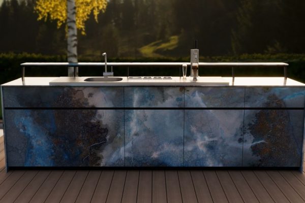 Theiss Outdoor-Bar mit blauen Steinfronten, Spülbecken und Zapfanlage auf Terrasse im Abendlicht mit Wald- und Bergkulisse.