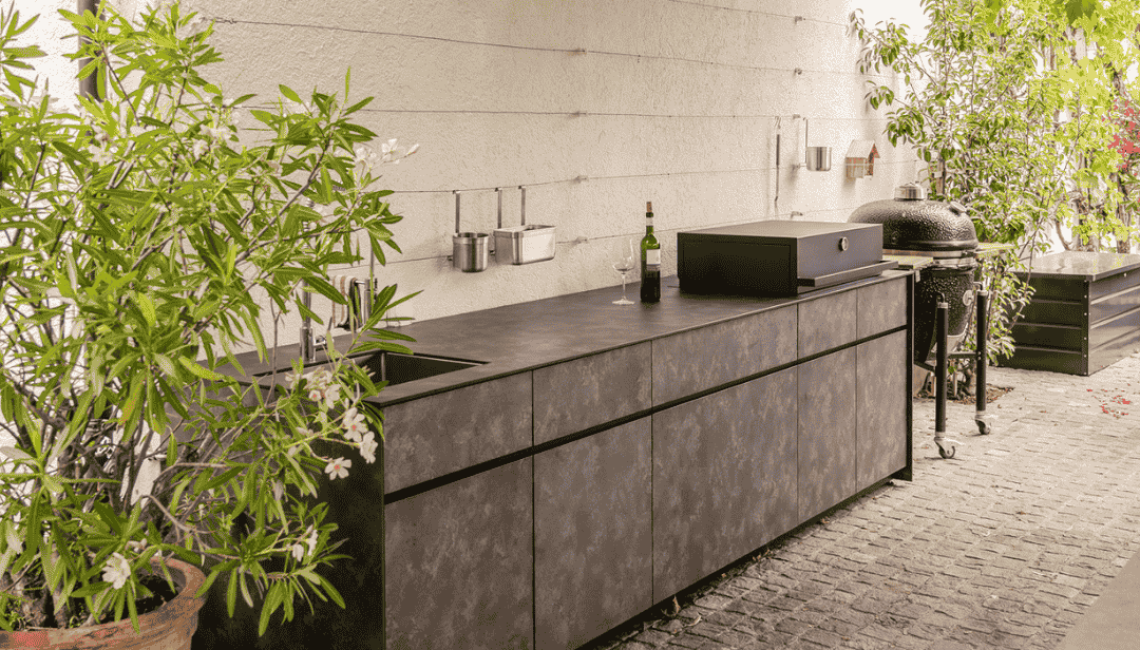 Aussenküche Theiss Avers in Stäfa mit HPL Stone ME Oberfläche, integriertem Spülbereich und Grillmodul entlang einer begrünten Hauswand