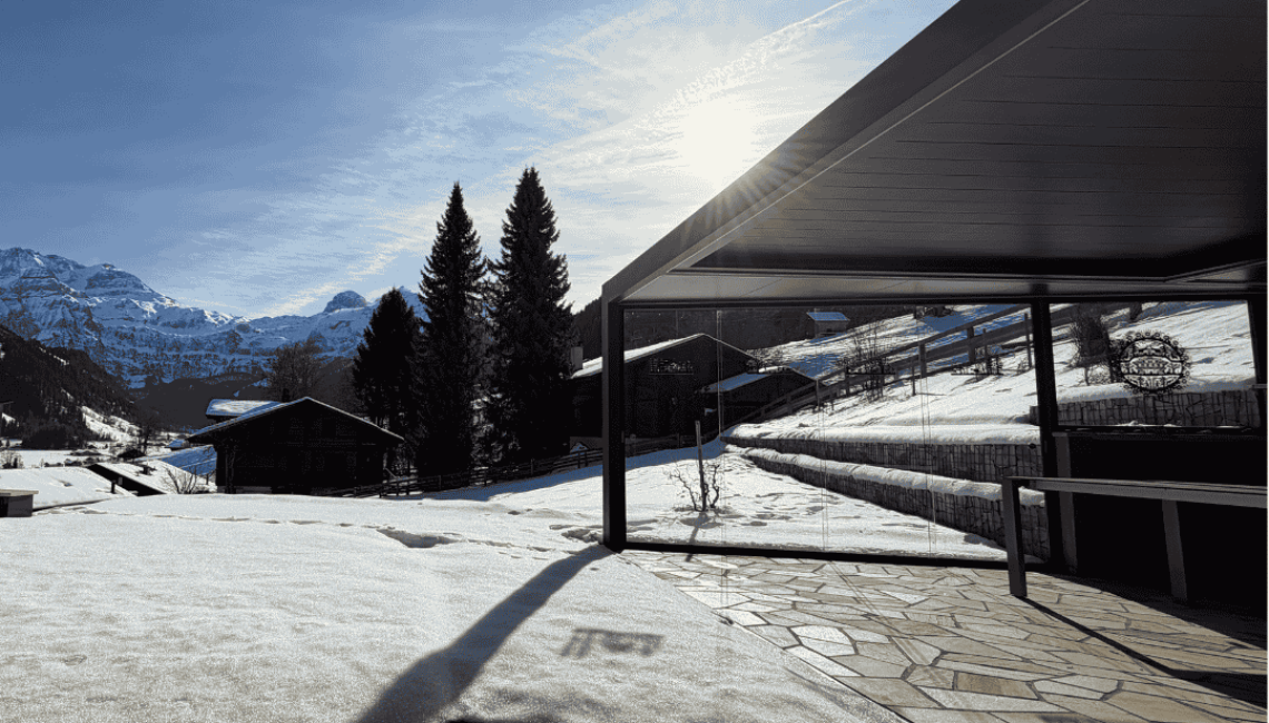 Lamellendach in Sonderform mit Ganz-Glas-Schiebeanlage auf einer Terrasse in Lenk im Simmental bei winterlichen Bedingungen mit Schnee und Bergpanorama.
