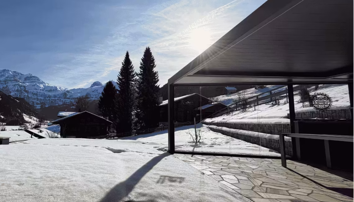 Lamellendach in Sonderform mit Ganz-Glas-Schiebeanlage auf einer Terrasse in Lenk im Simmental bei winterlichen Bedingungen mit Schnee und Bergpanorama.