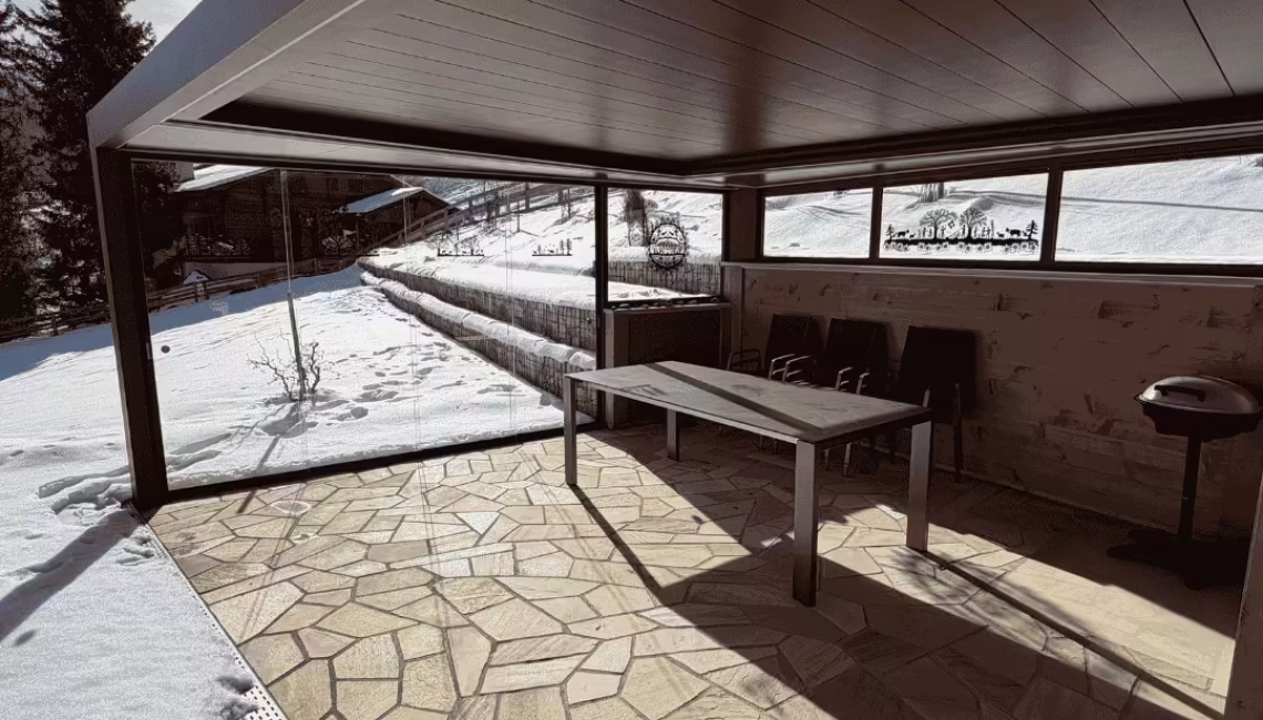 Überdachter Terrassenbereich mit Lamellendach und Ganz-Glas-Schiebeanlage in Lenk im Simmental, mit Sitzplatz, Tisch und winterlicher Umgebung.