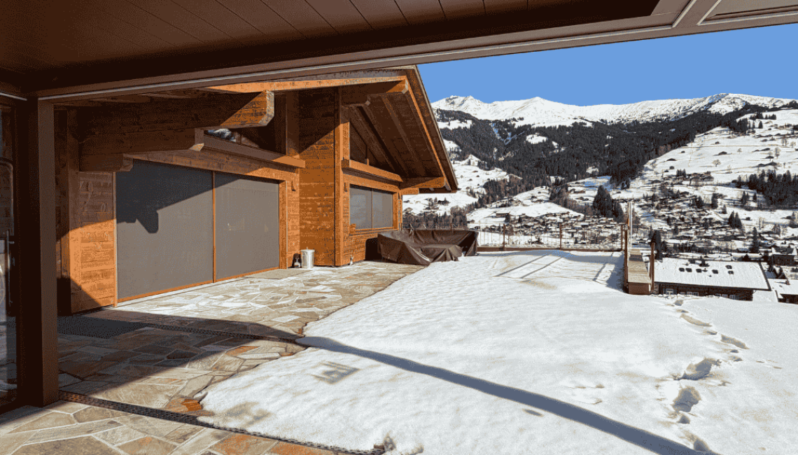 Blick von unter dem Lamellendach auf die verschneite Terrasse und die umliegende Berglandschaft in Lenk im Simmental.