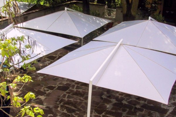 Weisse RIALTO Sonnenschirme auf einer gepflasterten Terrasse, die angenehmen Schatten unter Bäumen spenden.