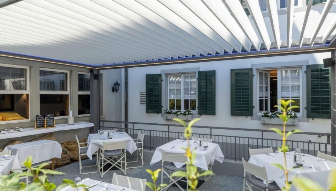 Restaurantterrasse in Küsnacht mit modernem Lamellendach, gedeckten Tischen und Blick auf die historischen Fenster mit grünen Läden.