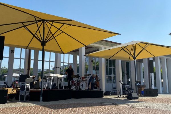 Zwei gelbe SCHATTELLO Grossschirme spenden Schatten auf einer Bühne mit Musikern bei einem Open-Air-Event.