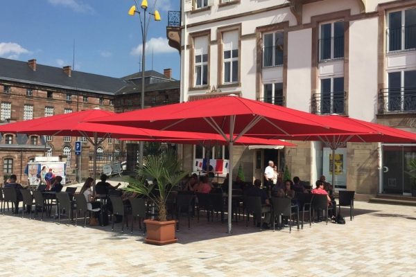 Zwei grosse rote SCHATTELLO Sonnenschirme spenden Schatten auf einer belebten Restaurantterrasse vor einem historischen Gebäude.