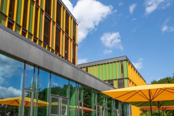 Gelbe SCHATTELLO Grossschirme spenden Schatten vor einem modernen Büro- und Institutsgebäude mit farbenfroher Architektur.