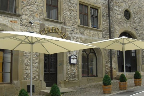 Helle SCHATTELLO Grossschirme vor einem historischen Gebäude, die den Aussenbereich eines Restaurants beschatten.