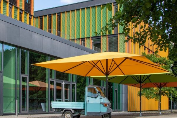 Mehrere farbige SCHATTELLO Grossschirme in Gelb, Grün und Orange vor einem modernen Gebäude mit auffälliger Architektur.