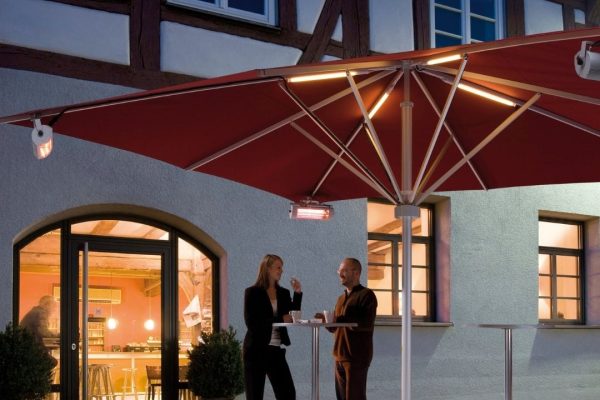Roter SCHATTELLO Grossschirm mit integrierter Beleuchtung und Heizstrahlern, unter dem zwei Personen an Stehtischen gemütlich im Freien stehen.