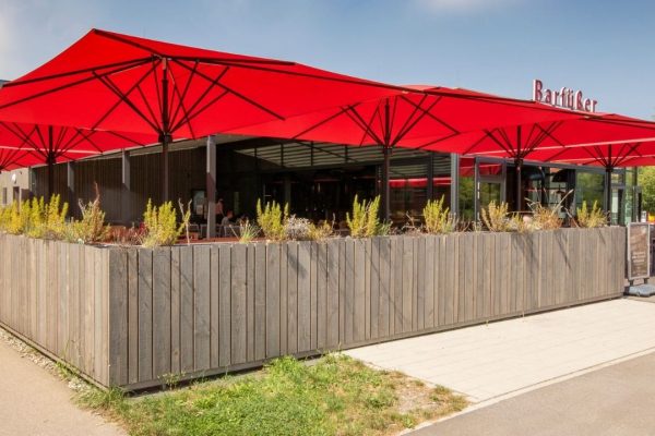 Rote SCHATTELLO Grossschirme spenden Schatten auf der Terrasse eines Restaurants mit moderner Holzverkleidung.