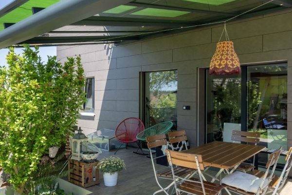 SGL 1 Glasdach mit grünem Sonnenschutz über einer gemütlichen Terrasse, geprüft bis 350 kg/m² Schneelast, moderne Lösung für Aussenbereiche.