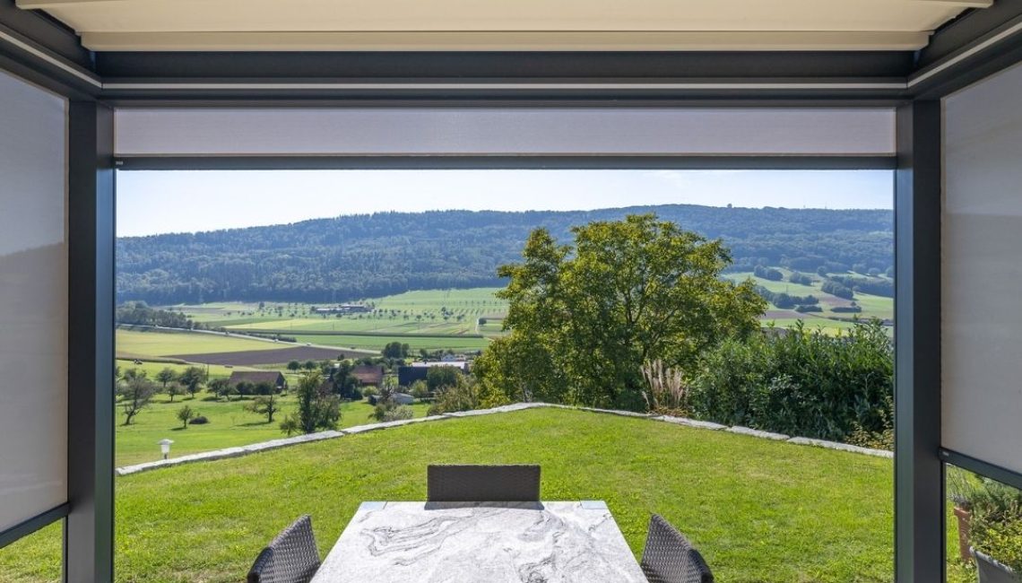 Textildach mit automatischen Senkrechtmarkisen und LED-Beleuchtung – moderner Essplatz mit Panoramablick auf Hügel und Felder.