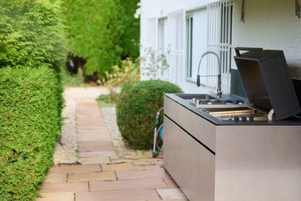 Moderne Valsot Outdoor-Küche mit integriertem Grill und Spülbecken in einer eleganten Natursteinoptik, platziert an einem Gartenweg.