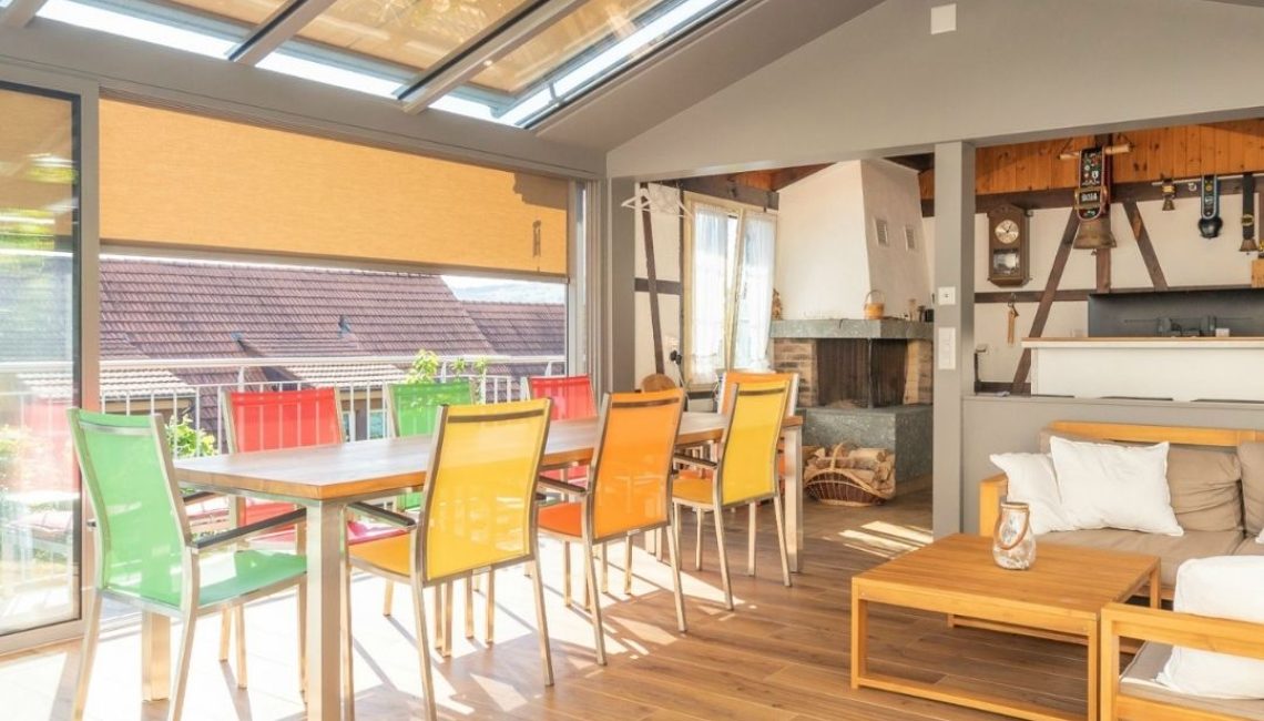 Wintergarten im Kanton Aargau mit Glasdach, Beschattung und Essbereich mit farbigen Stühlen.