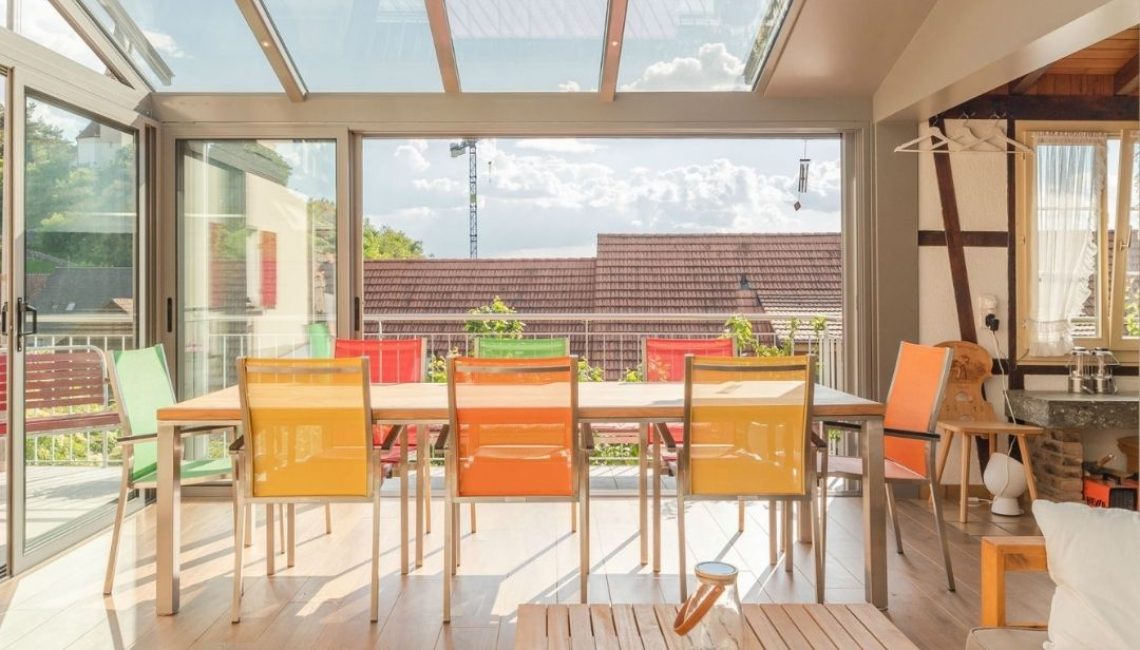 Wintergarten mit SGL1 Glasdach im Kanton Aargau, lichtdurchfluteter Essbereich mit bunten Stühlen und Ausblick ins Freie.