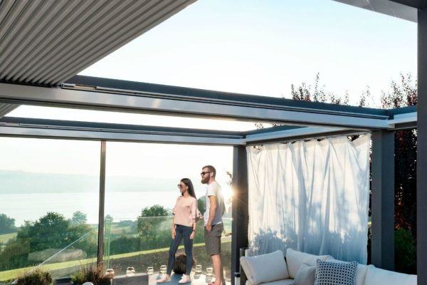 Panorama-Lamellendach mit Glasfront und Lounge, zwei Personen geniessen den Ausblick auf die Landschaft bei Sonnenuntergang.
